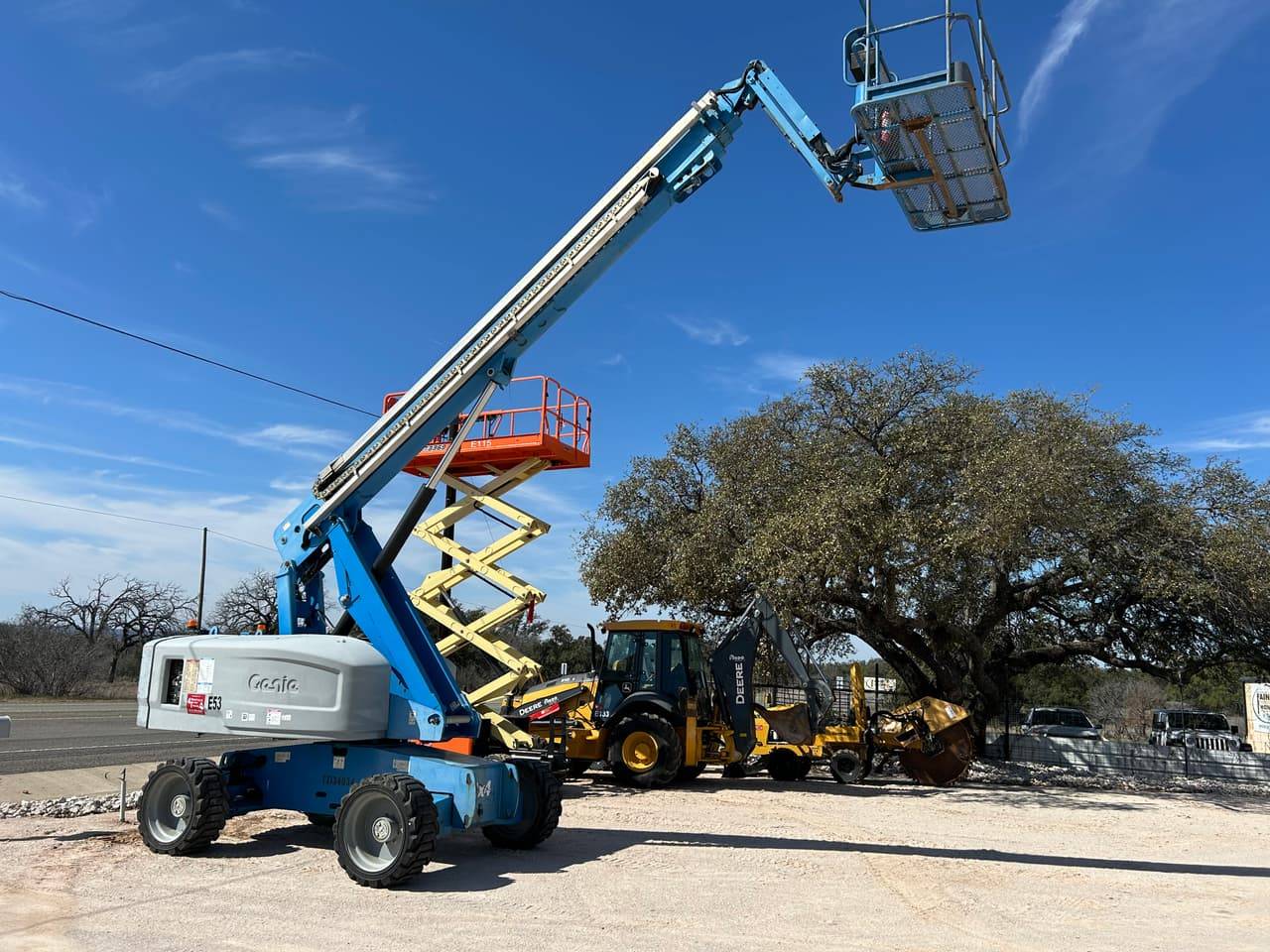 Genie boom lift with basket raised beside a JLG scissor lift and a John Deere backhoe on the 4-T Rental & Sales yard